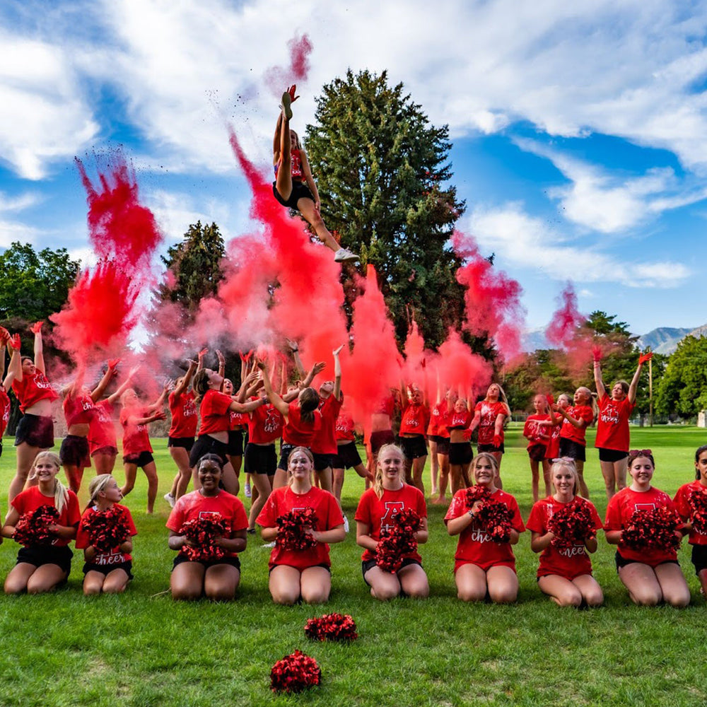 A group of cheerleaders in red uniforms celebrating with red smoke and pom-poms on a grassy field.