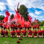A group of cheerleaders in red uniforms celebrating with red smoke and pom-poms on a grassy field.