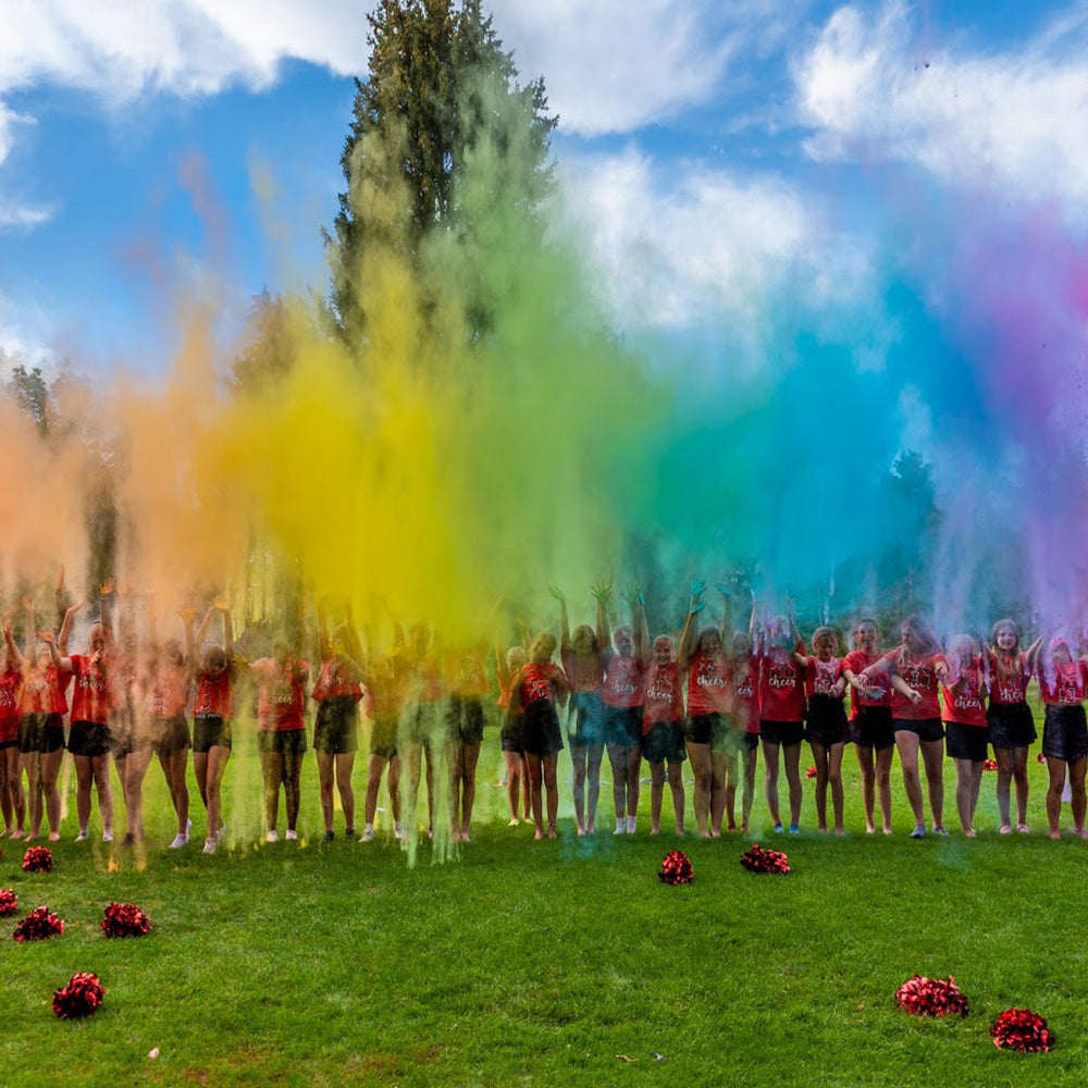 Cheerleaders in red shirts throw colorful powder, creating a rainbow cloud on a grassy field under blue sky. Pom-poms scattered. (68 characters)