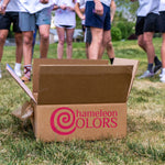 Open box with hameleon Colors logo on grass, surrounded by people in white shirts.