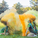 People in a grassy field throwing colorful powders, creating clouds of yellow, orange, and green during a festive event.