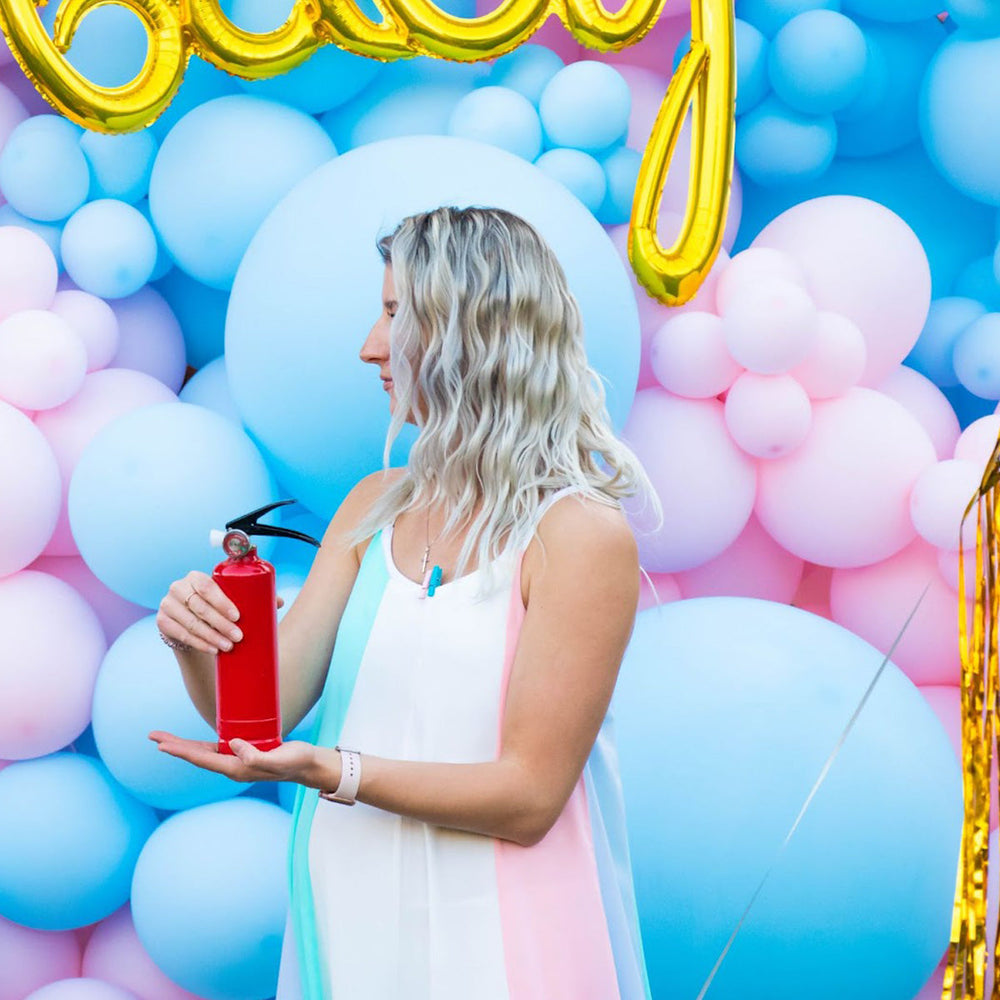 Pregnant woman at gender reveal party holds red fire extinguisher amid pink and blue balloons with Baby sign.