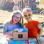 Two kids covered in colorful powder, smiling and giving thumbs up next to a box of Color Balls.