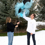 A vibrant gender reveal party outdoors with a family celebrating by popping a balloon filled with colorful powders.