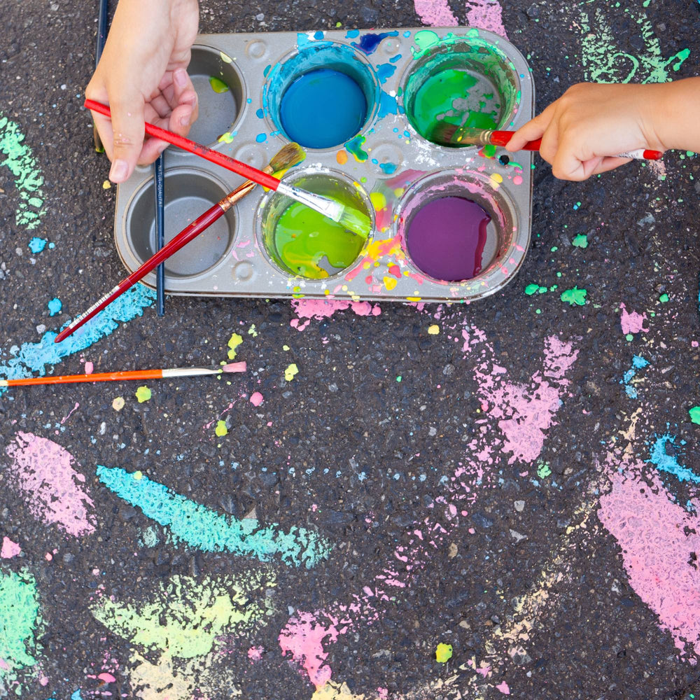 Two hands with paintbrushes dipping into a muffin tin filled with chalk paint on a pavement covered with strokes.